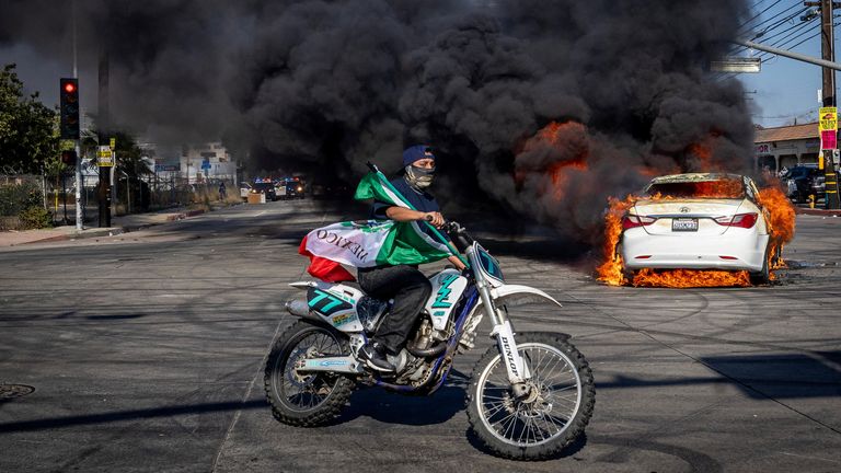 A man on a motorcycle carries a Mexican flag as smoke rises from a burning car on Atlantic Boulevard